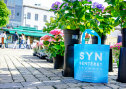 Synsenteret i Drammen er klar til høsten med nye poser! | Handlenett med trykk på en somemrlig Bragernes torg foran blomstre. | SKG - Spesialisten innen profilert emballasje