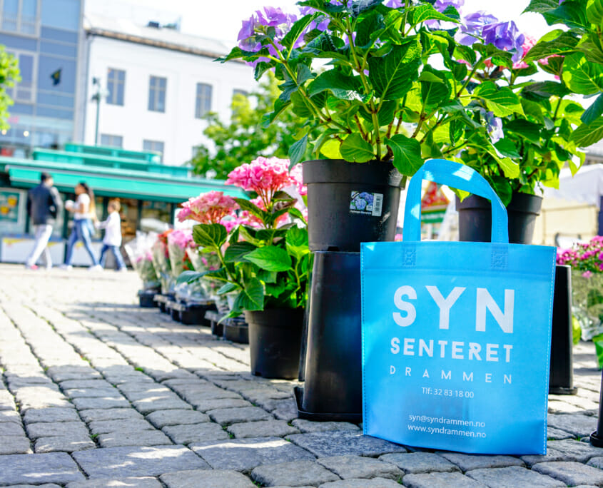 Synsenteret i Drammen er klar til høsten med nye poser! | Handlenett med trykk på en somemrlig Bragernes torg foran blomstre. | SKG - Spesialisten innen profilert emballasje