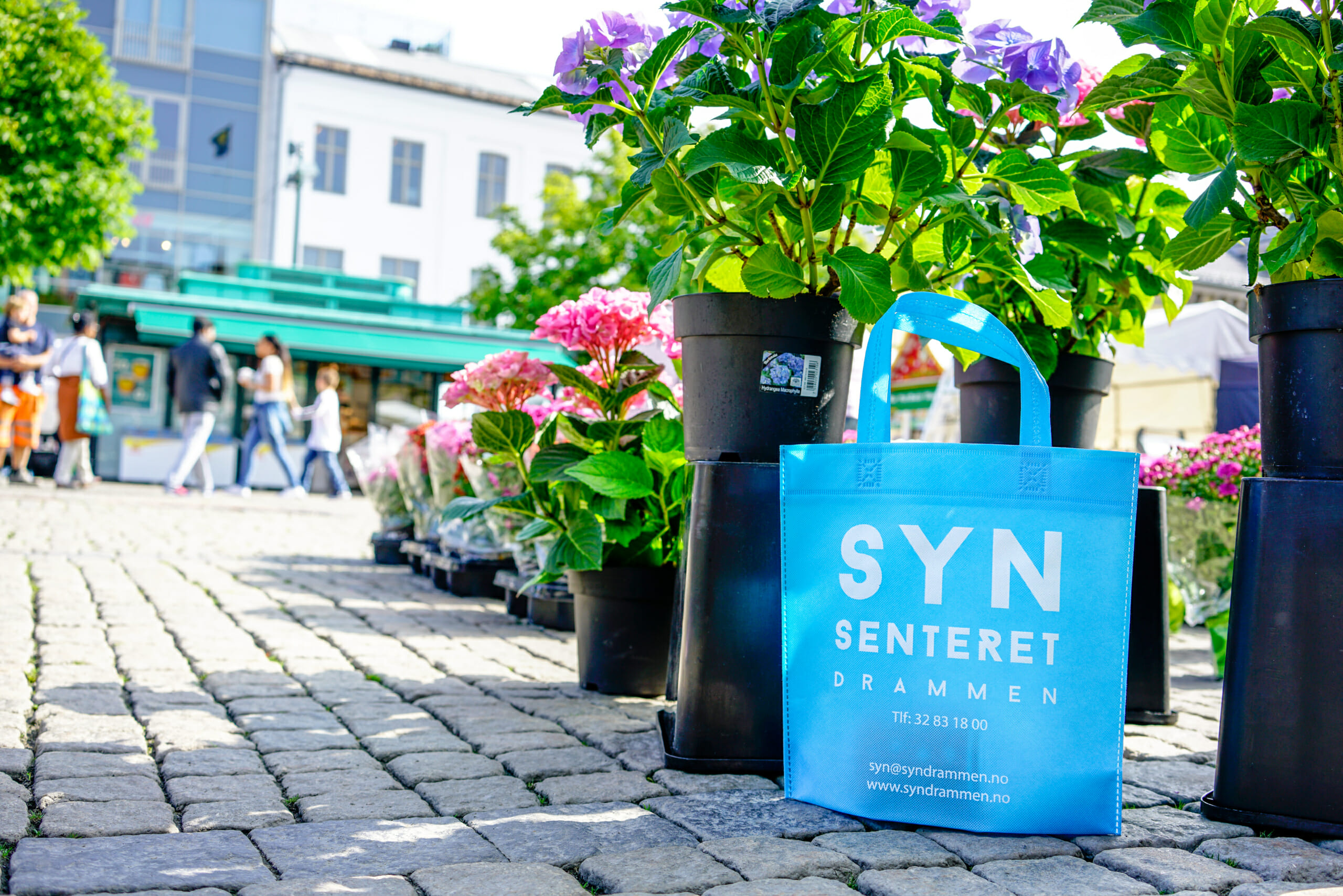 Synsenteret i Drammen er klar til høsten med nye poser! | Handlenett med trykk på en somemrlig Bragernes torg foran blomstre. | SKG - Spesialisten innen profilert emballasje